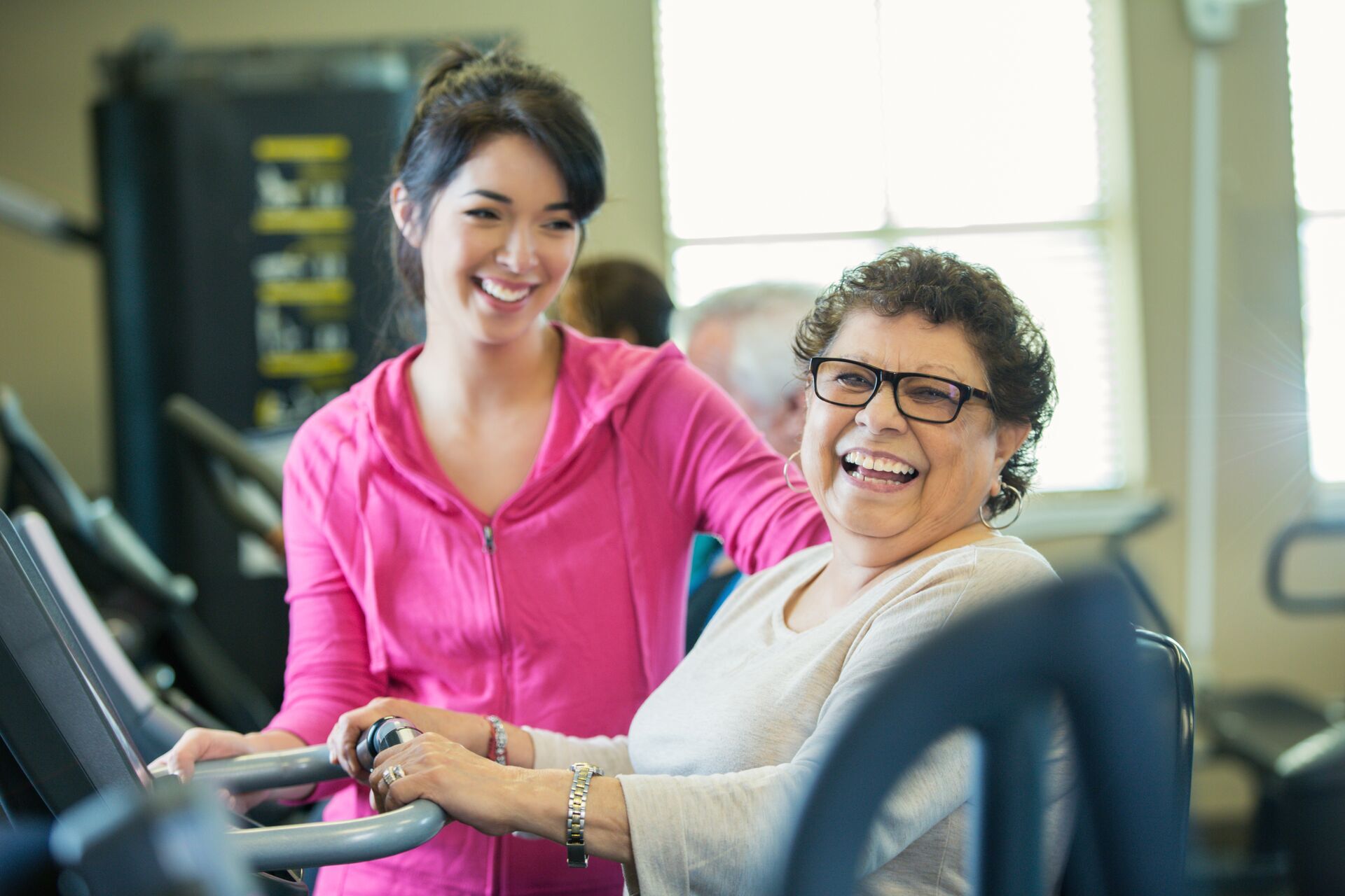 Beautiful Hispanic senior woman confidentlly uses machine at gym or senior center. Young female personal trainer helps the senior woman. They are both smiling. People are working out it the background.