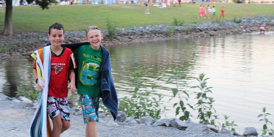 two boys smiling towards the camera with the YMCA Camp Thunderbird lake behind them and towels draped over their shoulders